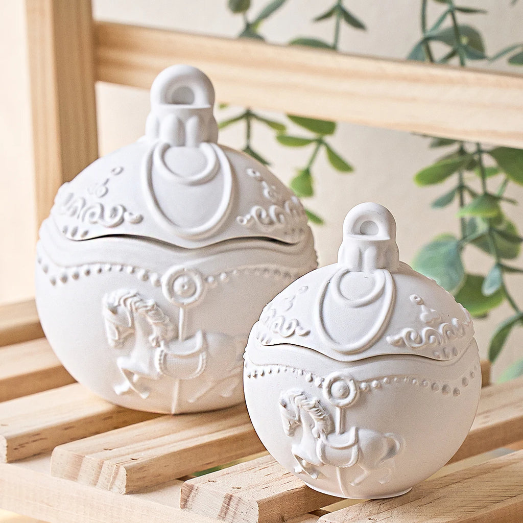 Two white ceramic decorative lidded containers with embossed carousel horse designs, displayed on a light wooden shelf with a soft-focus background featuring green leaves.
