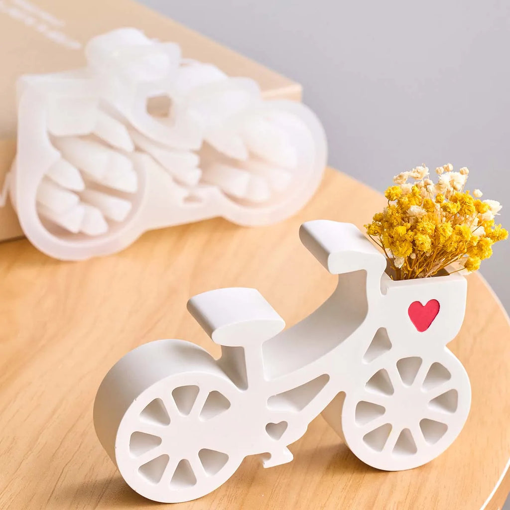A white bicycle-shaped decorative candle holder filled with yellow dried flowers, shown on a light wood surface with a blurred matching candle and box in the background.