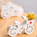 A white bicycle-shaped decorative candle holder filled with yellow dried flowers, shown on a light wood surface with a blurred matching candle and box in the background.