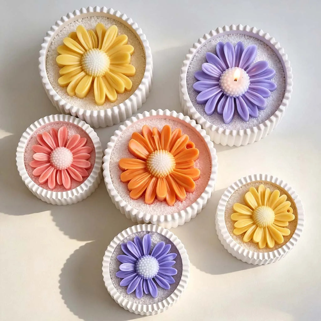 Seven small white cupcake-shaped items arranged on a light beige surface, each topped with a daisy flower in different colors including yellow, purple, pink, and orange.