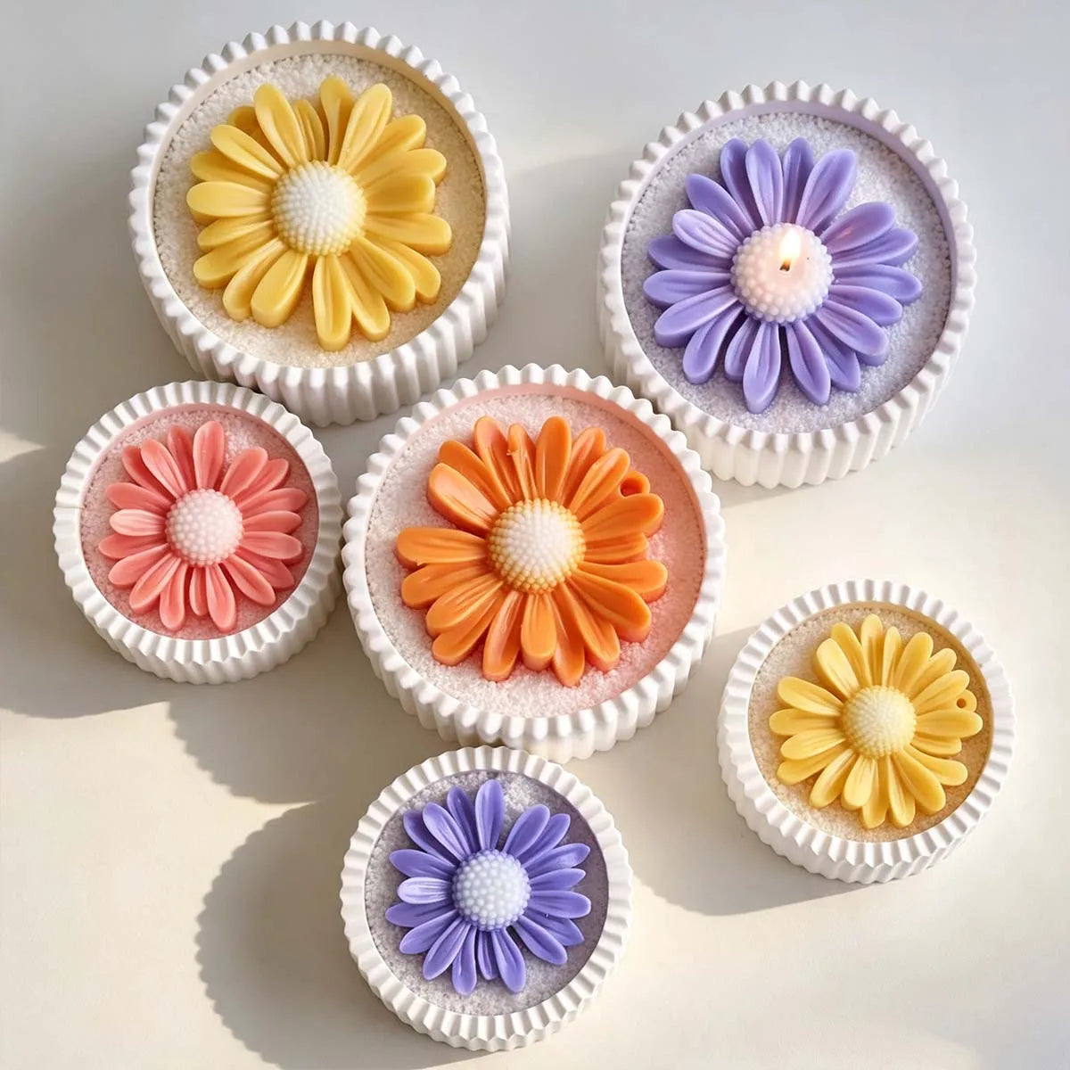 Seven small white cupcake-shaped items arranged on a light beige surface, each topped with a daisy flower in different colors including yellow, purple, pink, and orange.