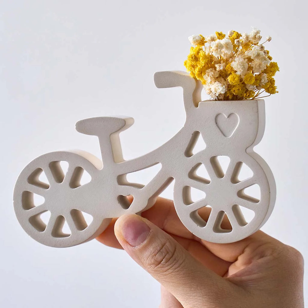 A hand holding a bicycle-shaped wooden planter with a small basket of yellow and white dried flowers, set against a plain light gray background.