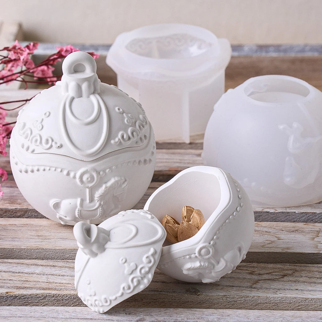 A white decorative ceramic ornament-shaped box with a hinged lid, shown open with a gold-toned ornament inside, alongside translucent white silicone mold halves on a rustic wooden surface.