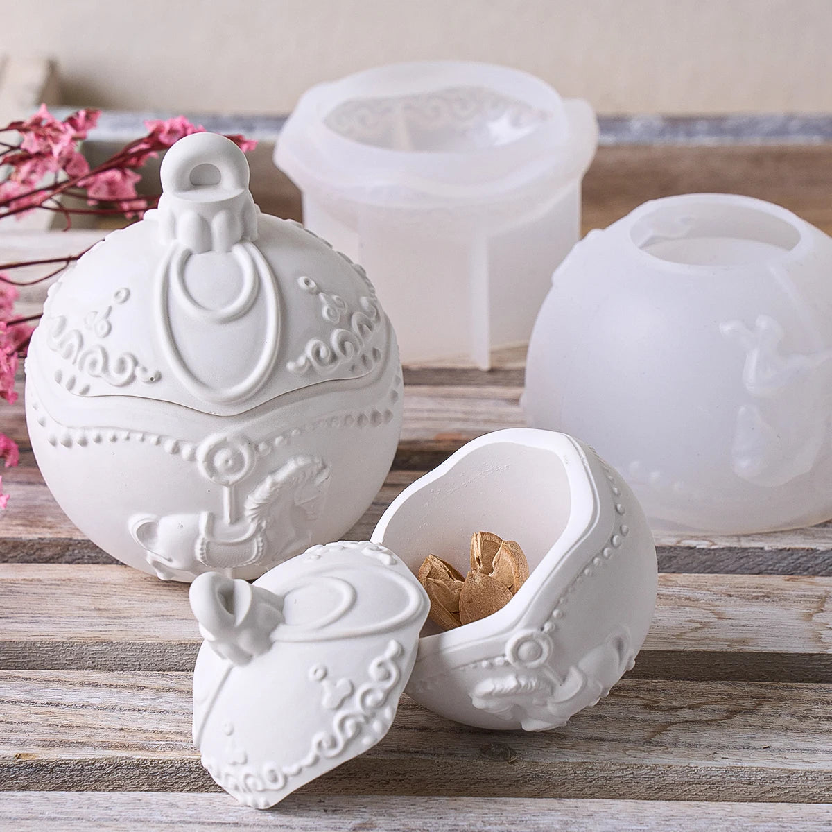 A white decorative ceramic ornament-shaped box with a hinged lid, shown open with a gold-toned ornament inside, alongside translucent white silicone mold halves on a rustic wooden surface.