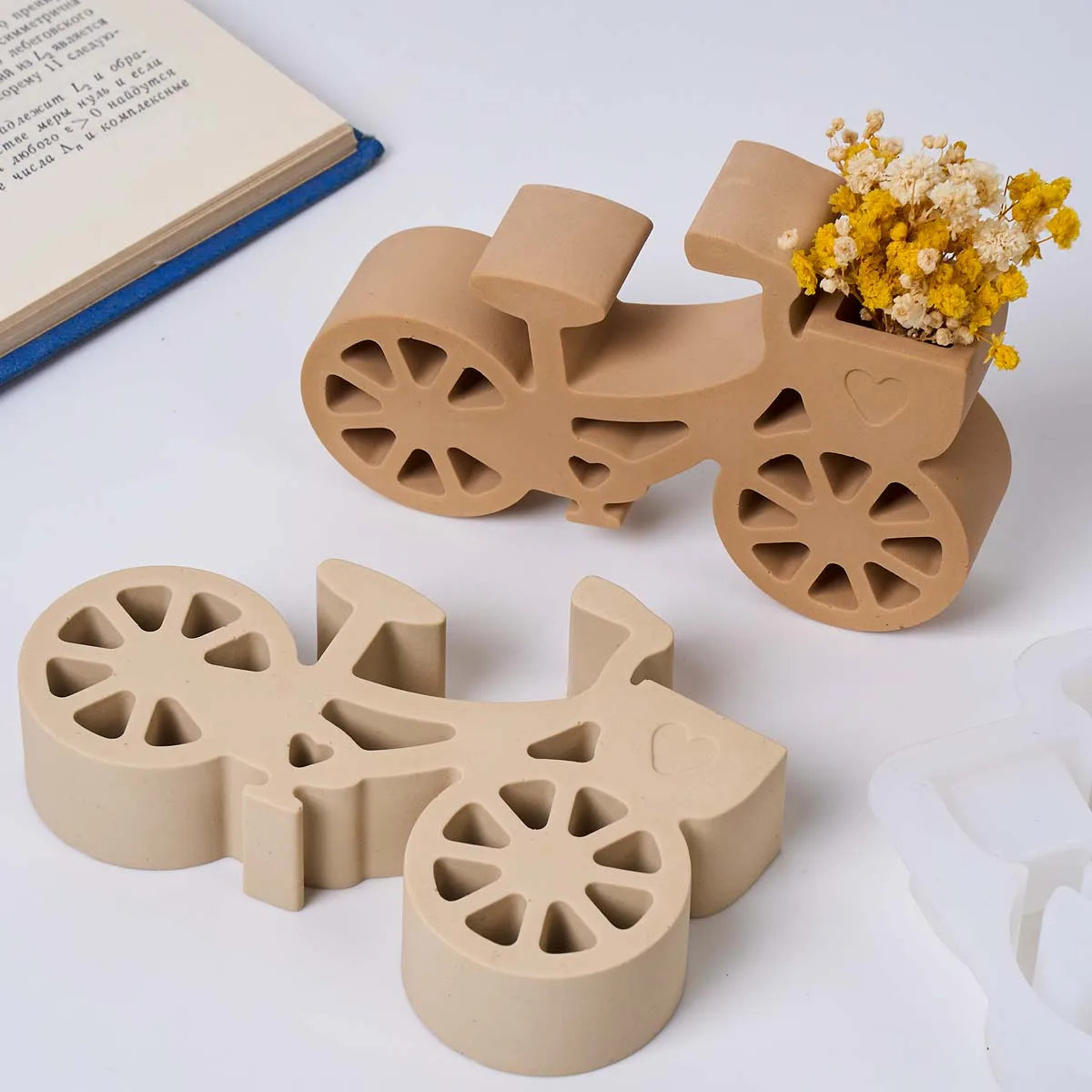 A bicycle-shaped decorative planter in a matte tan color, shown filled with dried yellow and white flowers on a white surface, alongside an empty matching planter and a white mold.