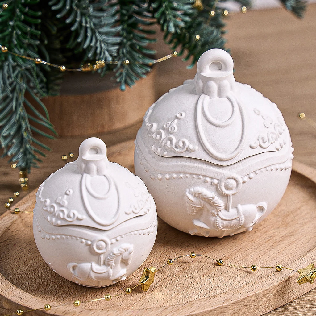 Two white ceramic Christmas ornament-shaped boxes with lids, featuring embossed scrollwork and carousel horse details, displayed on a wooden tray with holiday greenery and gold bead garlands.