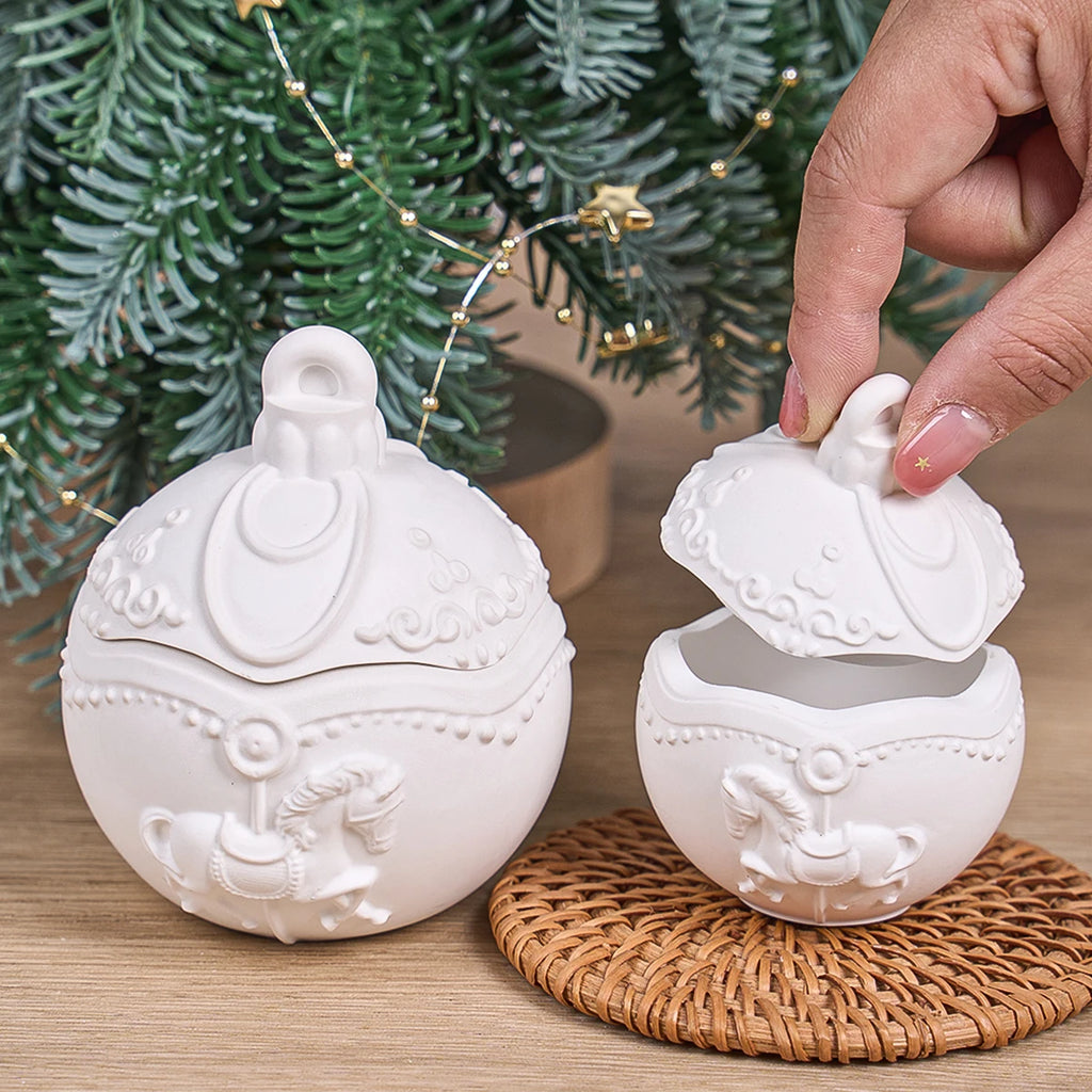 Two white ceramic Christmas ornament-shaped sugar bowls with lids, one sitting on a woven coaster and the other being lifted by a hand, set against a background of a green Christmas tree and warm string lights.