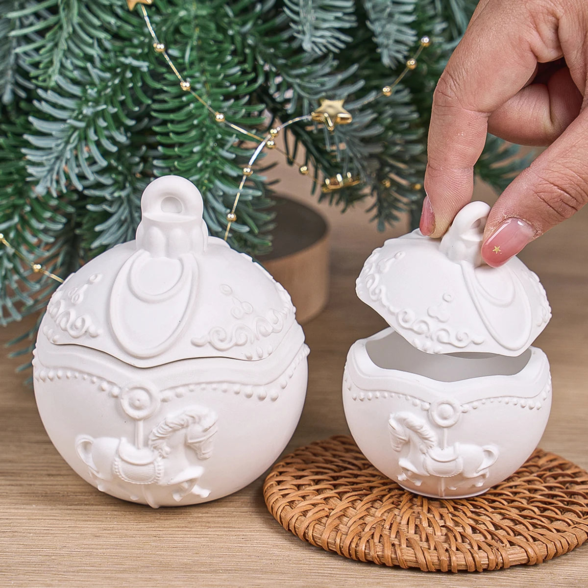 Two white ceramic Christmas ornament-shaped sugar bowls with lids, one sitting on a woven coaster and the other being lifted by a hand, set against a background of a green Christmas tree and warm string lights.