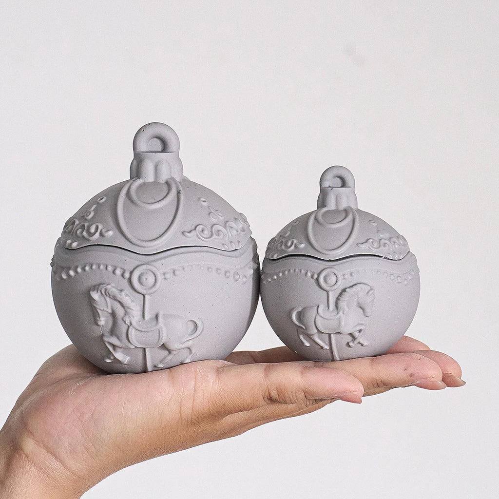 Two small round gray decorative lidded boxes with embossed carousel horse designs, held in a person's hand against a plain light background.