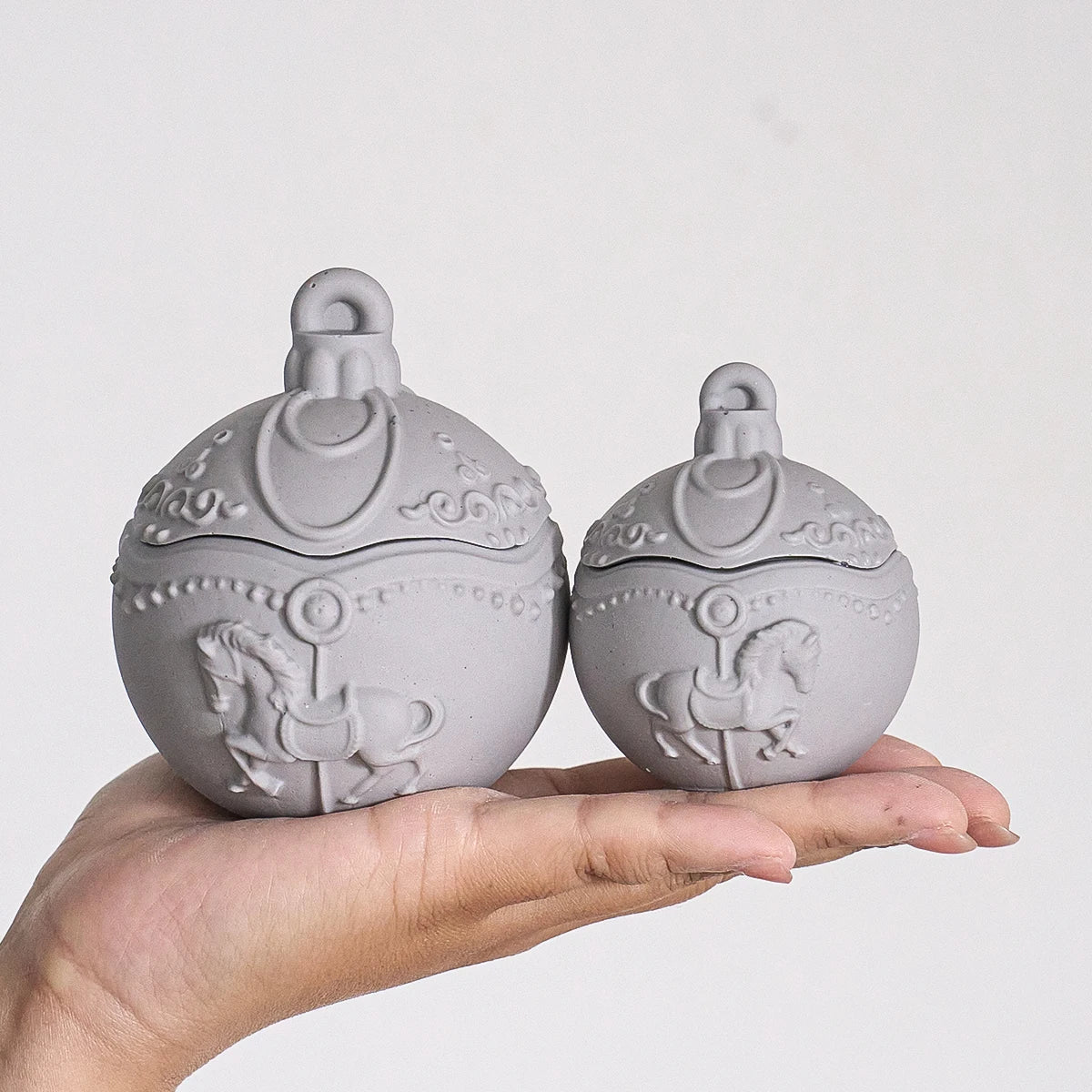 Two small round gray decorative lidded boxes with embossed carousel horse designs, held in a person's hand against a plain light background.