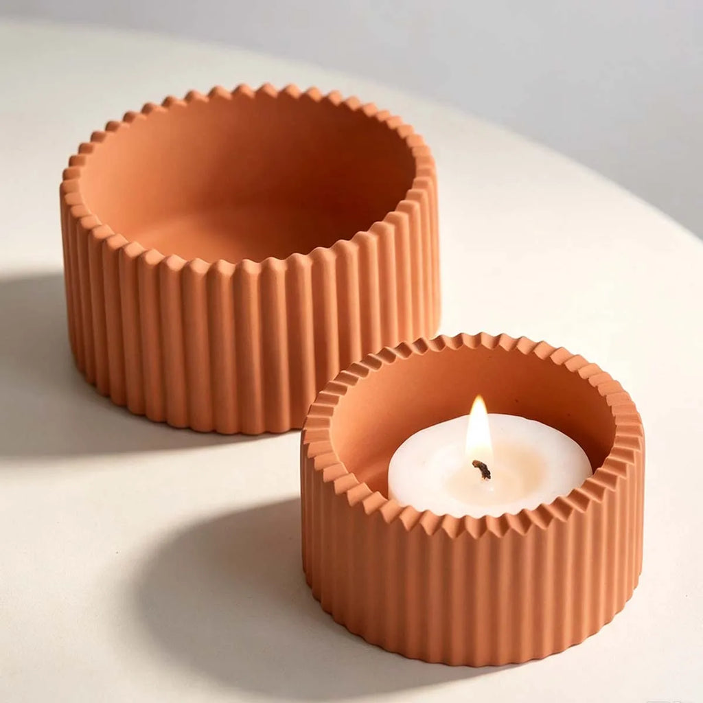 Two terracotta-colored ribbed candle holders on a light surface, with one holder lit and holding a white tealight candle.