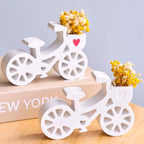 Two white bicycle-shaped ceramic vases filled with yellow dried flowers, displayed on a light wood surface with a beige book in the background.