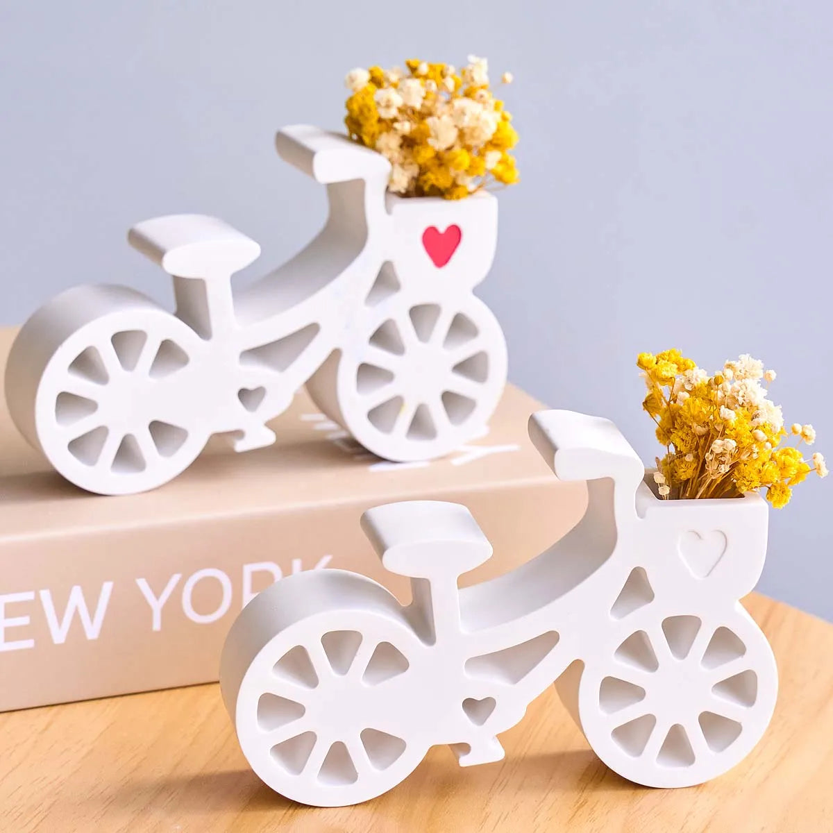 Two white bicycle-shaped ceramic vases filled with yellow dried flowers, displayed on a light wood surface with a beige book in the background.
