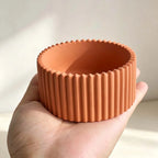 A terracotta-colored silicone cup with a fluted, ribbed exterior and scalloped rim, held in a person's hand against a soft, light beige background.