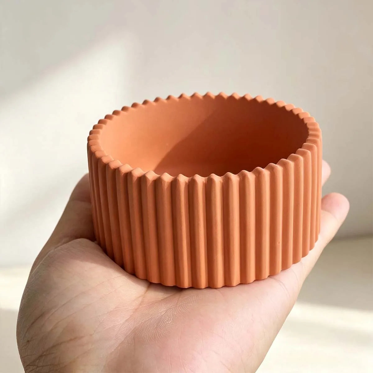A terracotta-colored silicone cup with a fluted, ribbed exterior and scalloped rim, held in a person's hand against a soft, light beige background.
