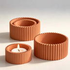 Three terracotta-colored ribbed candle holders on a light gray background, with one holder in the front left containing a lit white tea light candle.